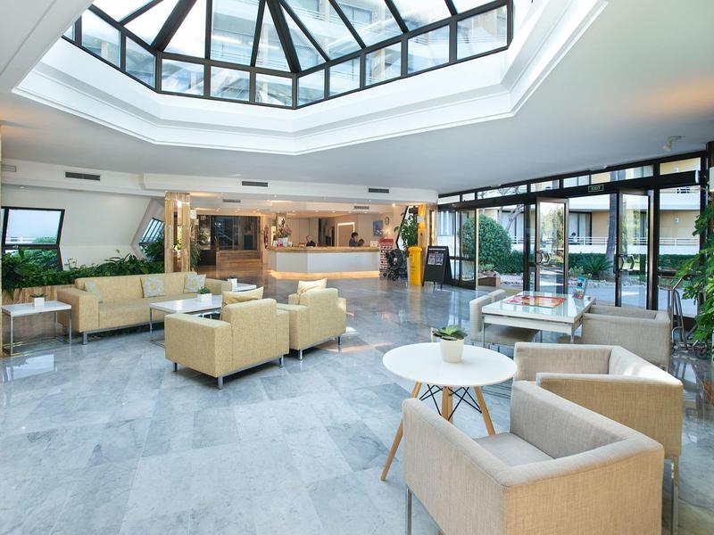 Heller, moderner Lobbybereich mit bequemen Sesseln, großen Fenstern und Glasdach für Tageslicht.