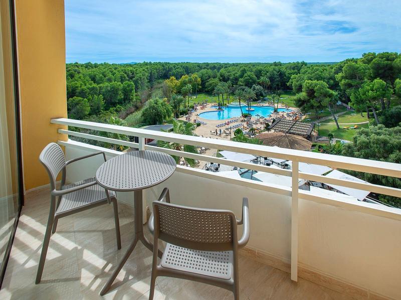 Balkon mit kleinem runden Tisch, zwei Stühlen und Blick auf Pool, grüne Bäume und blauen Himmel.