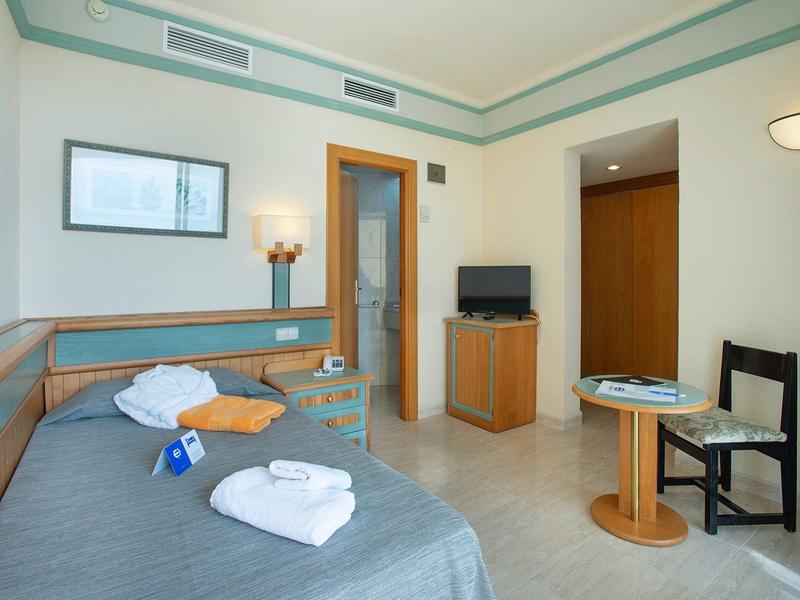 Hotelzimmer mit Einzelbett, Handtüchern, Tisch, Stuhl, Schrank und Lampen in hellem Raum.