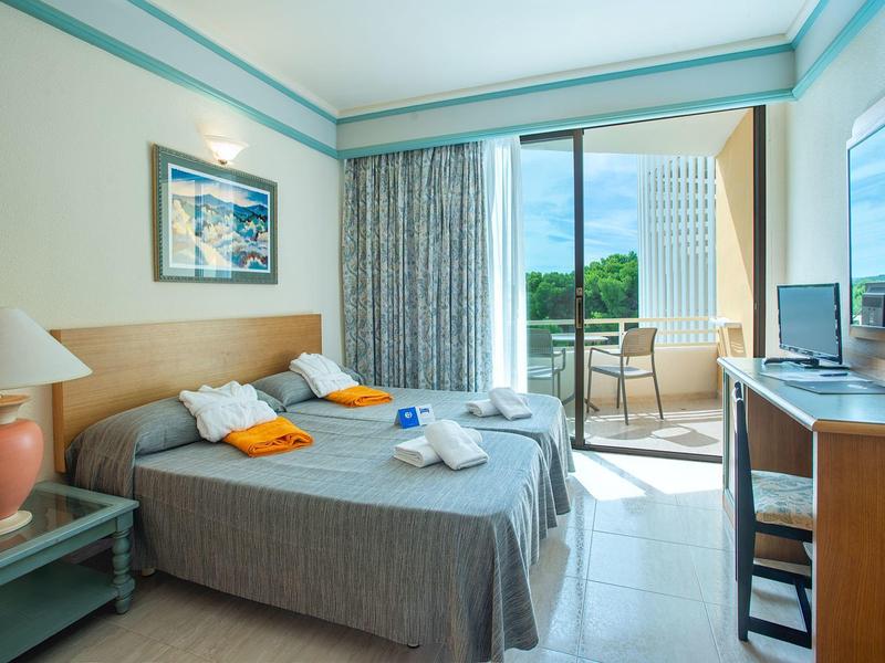 Helles Hotelzimmer mit zwei Einzelbetten, Schreibtisch, Fernseher und Balkon mit grünem Ausblick.