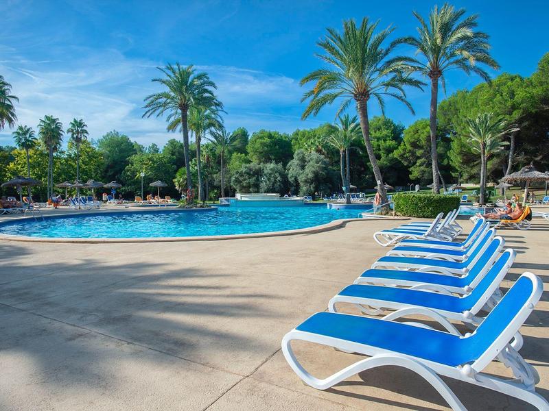Freier Pool mit blauen Liegestühlen, umgeben von Palmen und grünem Garten unter blauem Himmel.