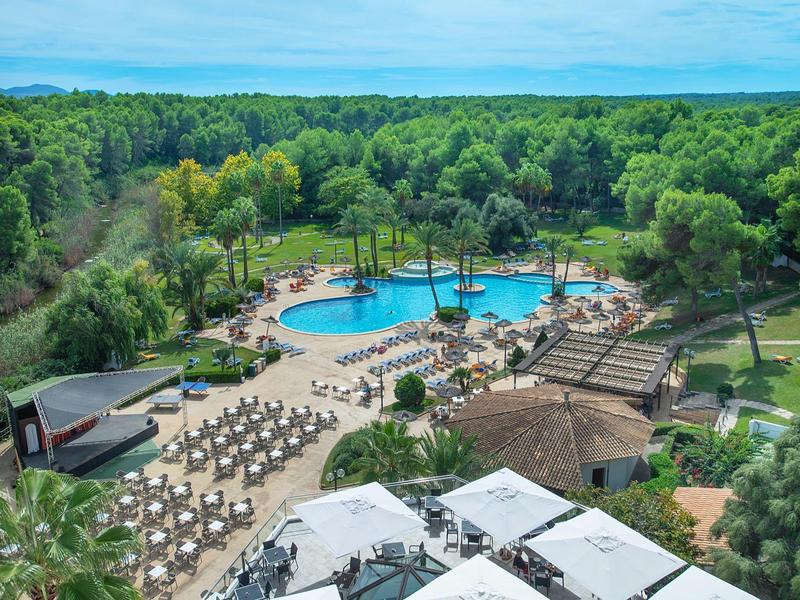 Großer Pool mit Sonnenschirmen und Liegen, umgeben von dichtem grünen Wald und blauem Himmel.
