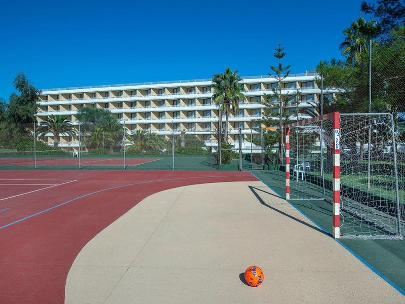 Leerer Handballplatz mit Tor, orangefarbener Ball, Palmen und mehrstöckigem Gebäude im Hintergrund.