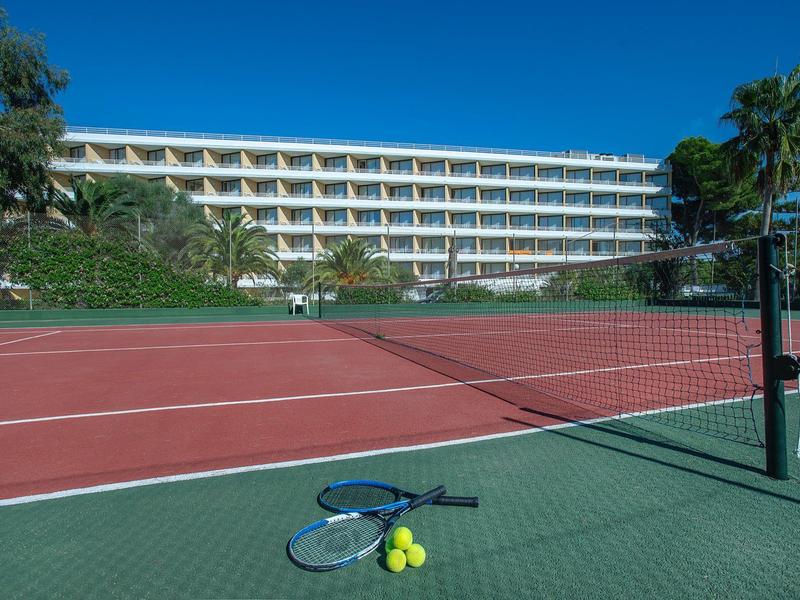 Roter Tennisplatz mit Netz, Tennisschläger und Bällen, im Hintergrund ein mehrstöckiges Hotel.