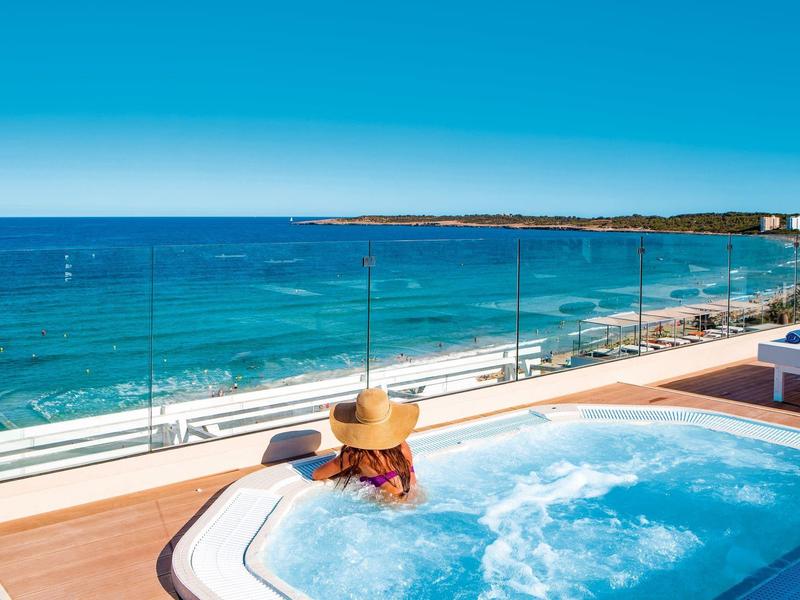 Person with sunhat relaxes in a hot tub on a terrace overlooking the sea and beach.