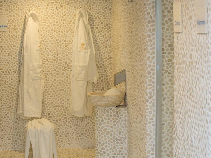 Two white bathrobes hang on the wall of a bathroom decorated with stone mosaic.