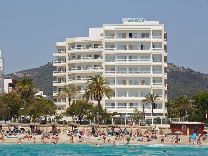 Hotel building on the beach with many sunbathers and clear blue water in the foreground.