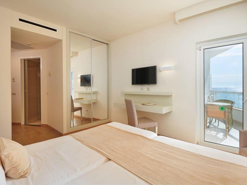 Bright hotel room with double bed, wall-mounted TV, and balcony access overlooking the sea.
