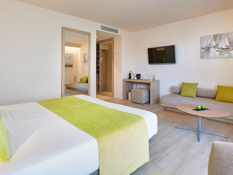 Modern hotel room with double bed, sofa, TV, and light wooden furniture.