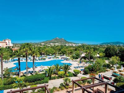 Pohled na hotelový bazén obklopený palmami s kopci v pozadí pod jasnou oblohou.