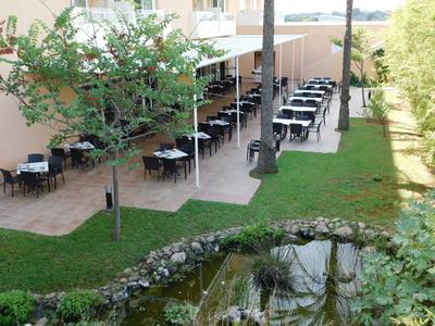 Venkovní prostor restaurace s stolky vedle malého rybníka a stromů.