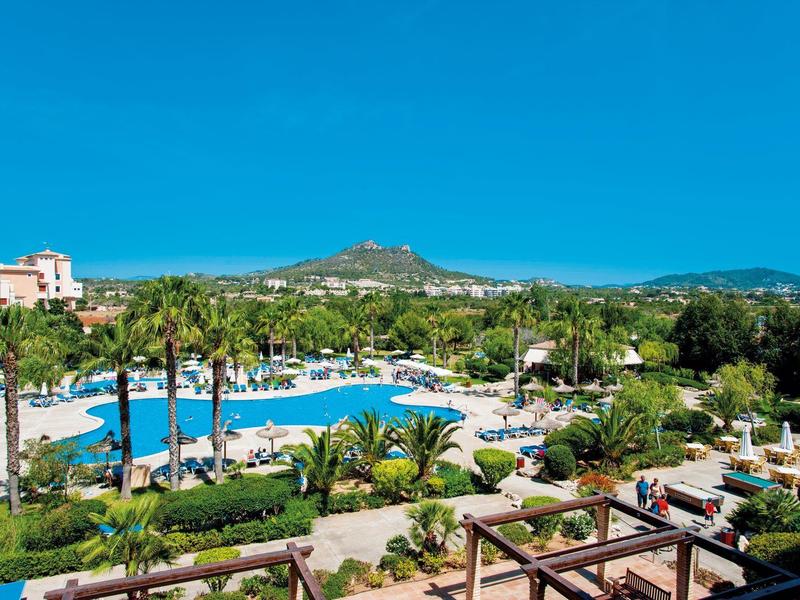 Pohled na hotelový bazén obklopený palmami s kopci v pozadí pod jasnou oblohou.