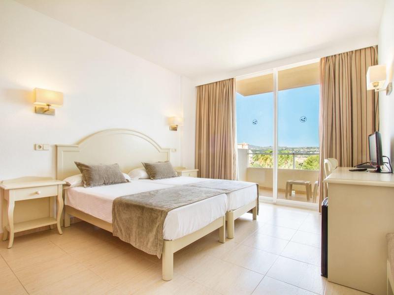 Světly hotelový pokoj s dvojlůžkem a balkonem s výhledem na moře.