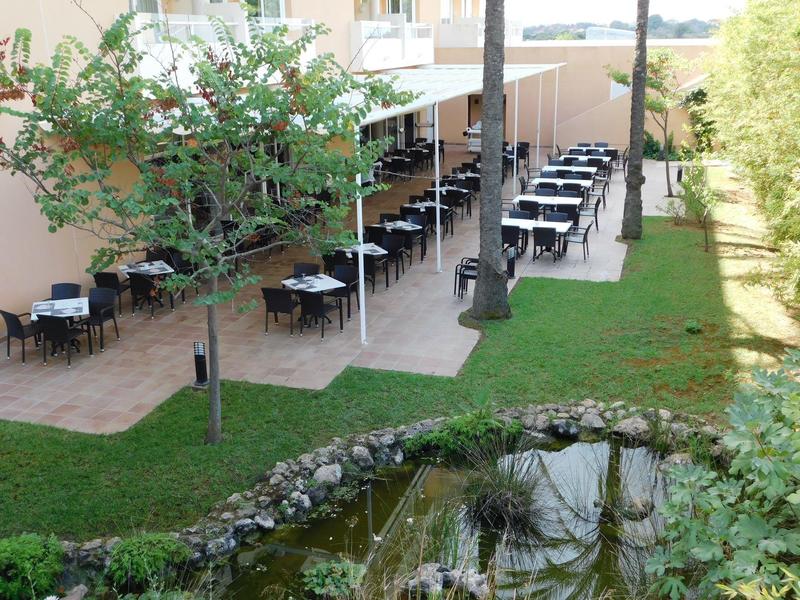 Venkovní prostor restaurace s stolky vedle malého rybníka a stromů.