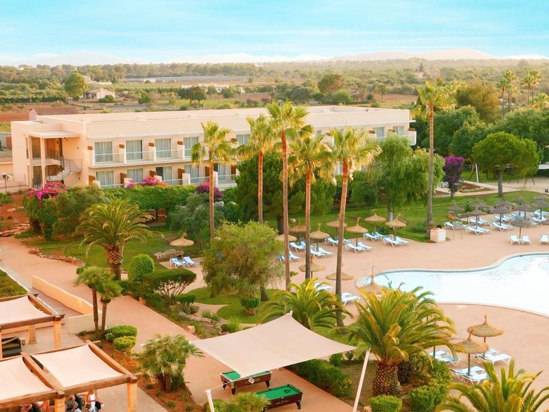 Hotel s bazénem, palmami a zahradou pod modrou oblohou.