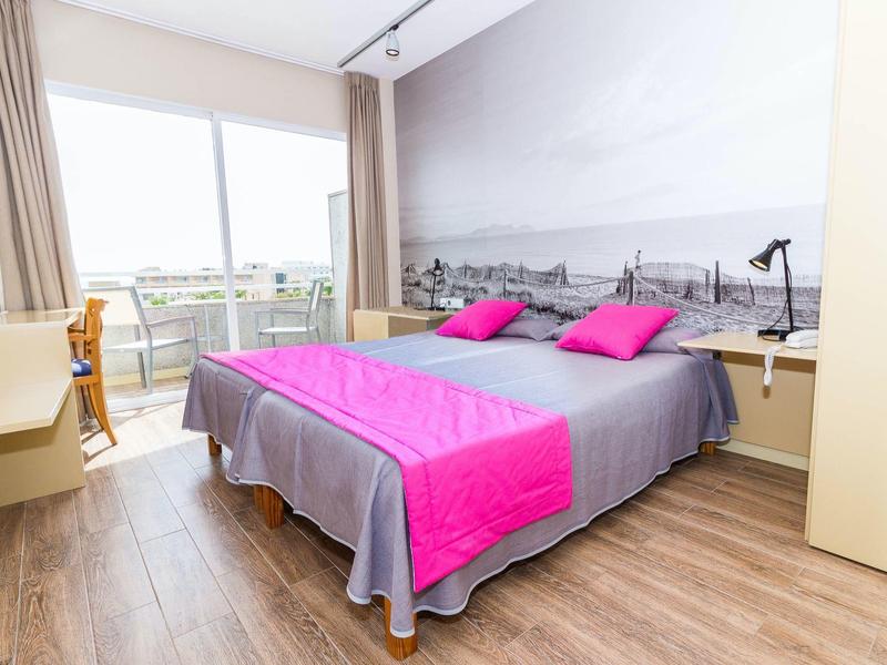 Bright hotel room with double bed, pink bedding, and large window with outdoor view.
