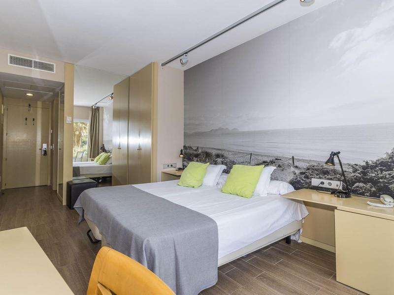 Modern hotel room with double bed, large wall mural, and simple decor in neutral tones.