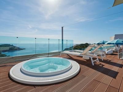 Jacuzzi auf Holzterrasse mit Blick auf das Meer und Liegestühle bei sonnigem Himmel.