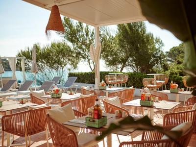 Restaurant terrace with tables, chairs, and view of pool and greenery in daylight.