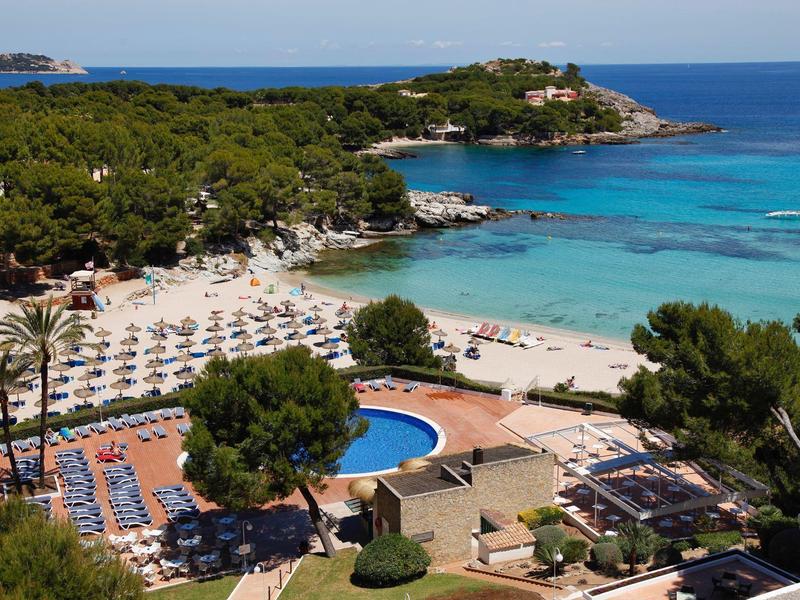 Station balnéaire avec parasols, piscine et mer turquoise entourée de verdure.