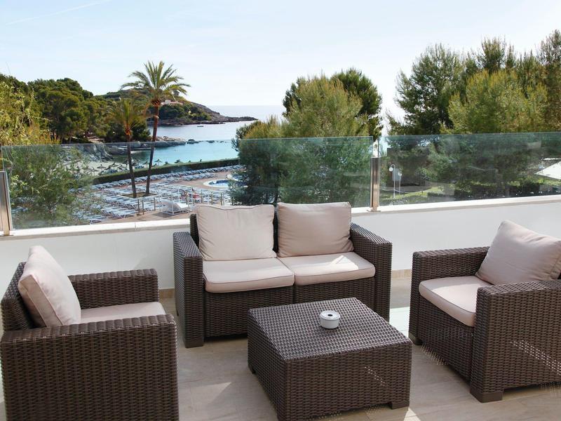 Coin salon extérieur avec mobilier en osier donnant sur un jardin et la mer au-delà d'une balustrade en verre.
