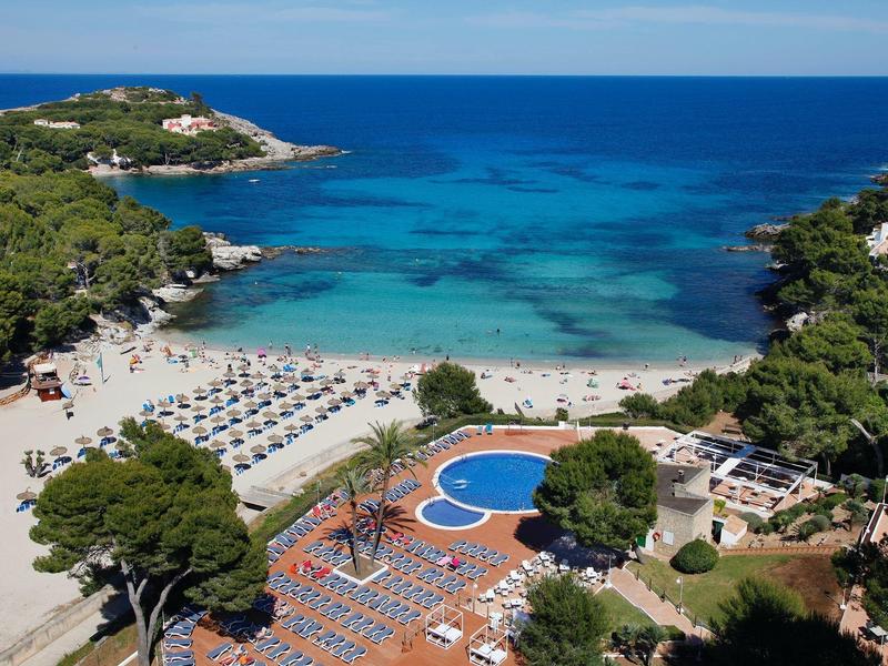 Plage ensoleillée avec parasols, sable, mer bleue et piscine entourée d'arbres.