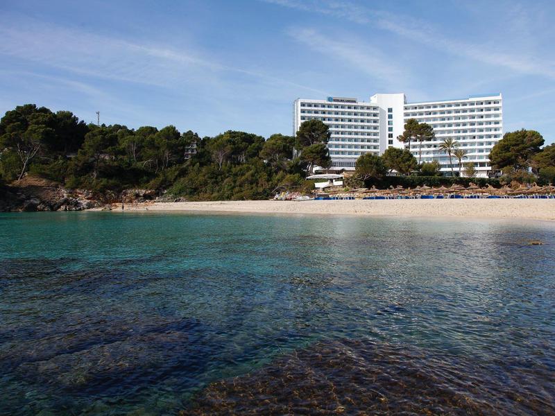 Hôtel au bord de la plage avec une eau bleue claire et des arbres sous un ciel partiellement nuageux.