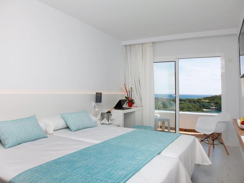 Chambre d'hôtel lumineuse avec lit double, accents turquoise, bureau, et porte vitrée donnant sur la mer