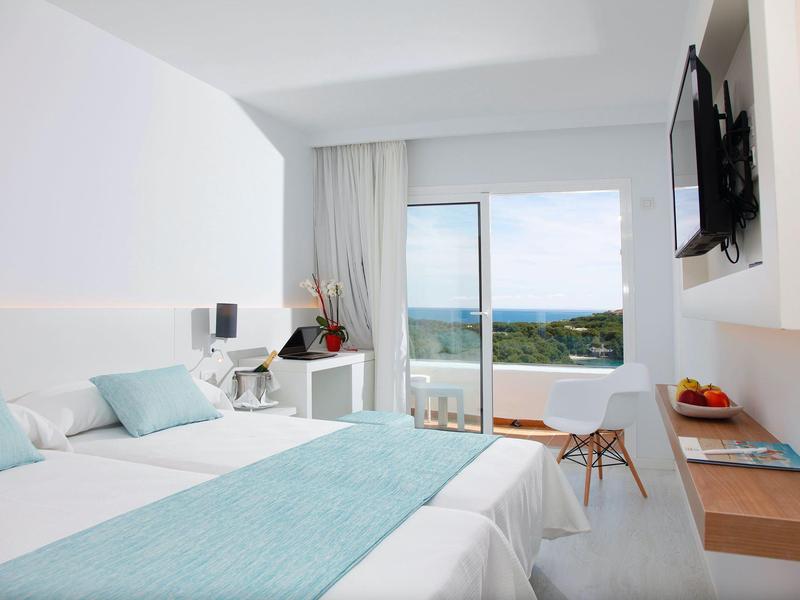 Chambre d'hôtel lumineuse avec lit double, accents turquoise, bureau et balcon avec vue sur l'océan.