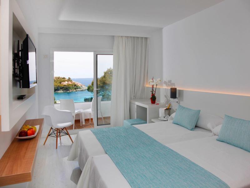 Chambre d'hôtel lumineuse avec deux lits, accents bleus et balcon donnant sur la mer.