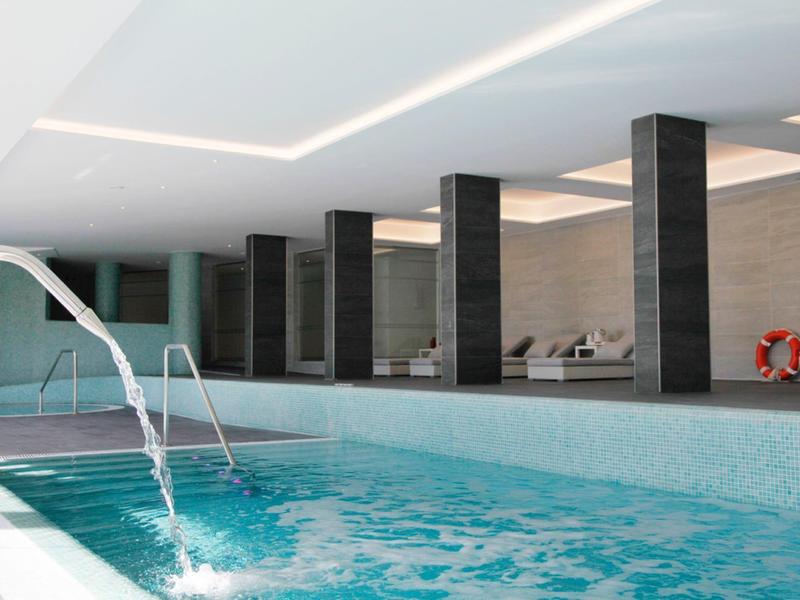 Piscine intérieure avec jets d'eau, murs carrelés et chaises longues dans un spa d'hôtel moderne.