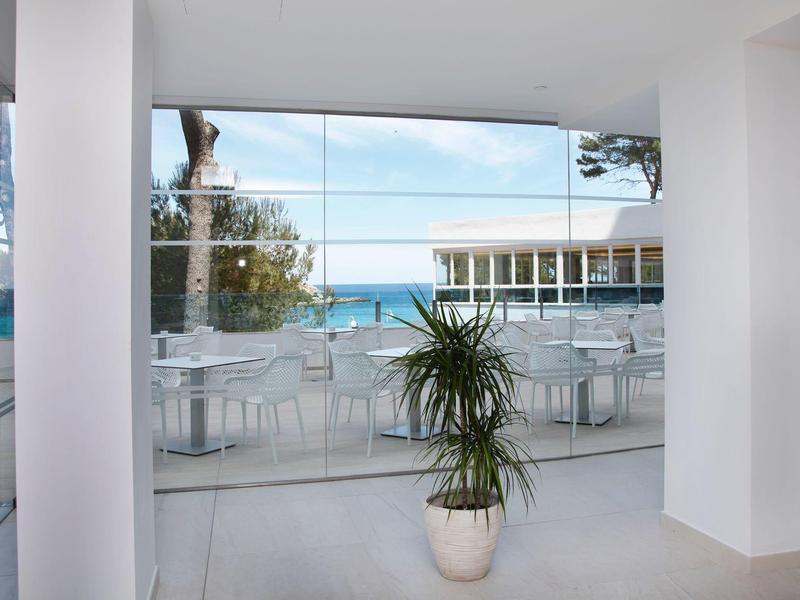 Terrasse d'hôtel lumineuse avec tables blanches, chaises et plante en pot face à la mer.