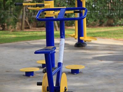 Blaue und gelbe Outdoor-Fitnessgeräte in einem Park mit Bäumen und Gebäuden im Hintergrund
