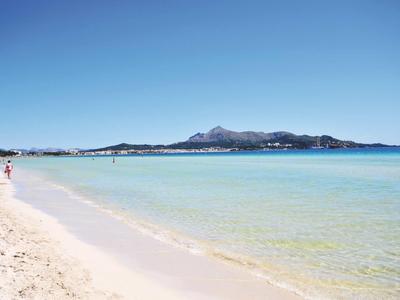 Jasná písčitá pláž s klidnou tyrkysovou vodou a horami v pozadí pod modrou oblohou.