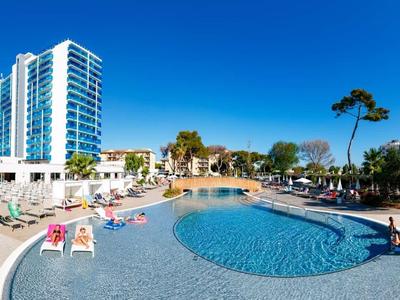 Kruhový bazén s lehátky, palmami a vysokou budovou pod modrou oblohou.