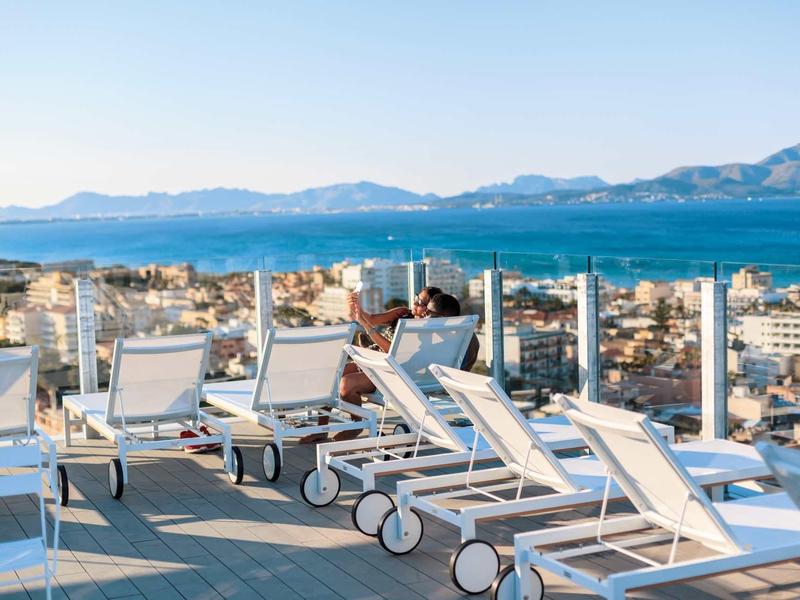 Prázdná lehátka na terase s výhledem na moře a hory v pozadí.