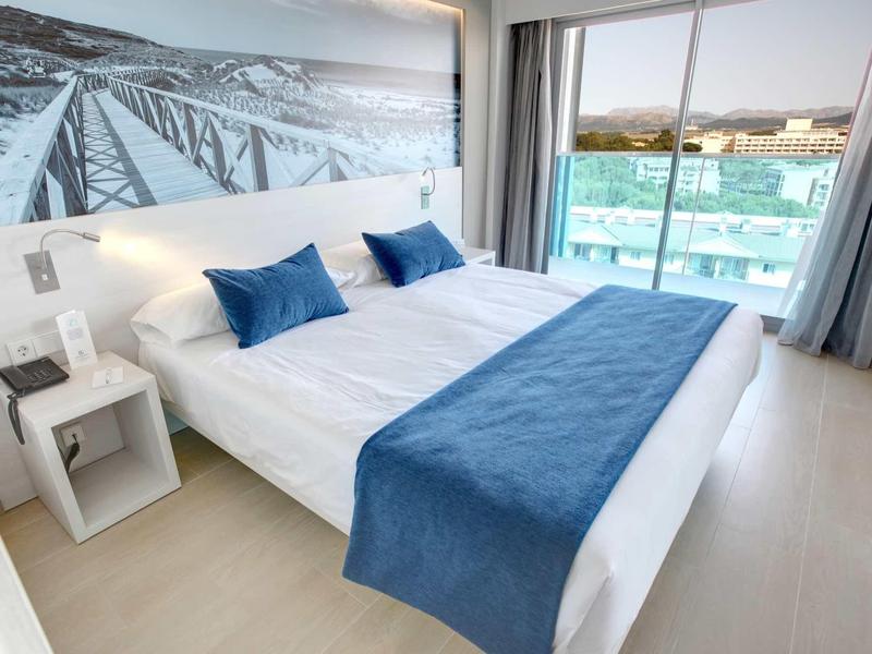 Moderní hotelový pokoj s manželskou postelí, modrým přehozem, polštáři a terasou s výhledem na moře.