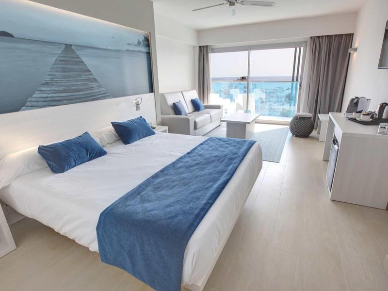 Světý hotelový pokoj s dvojlůžkem, modrým přehozem a balkonem s výhledem na moře.