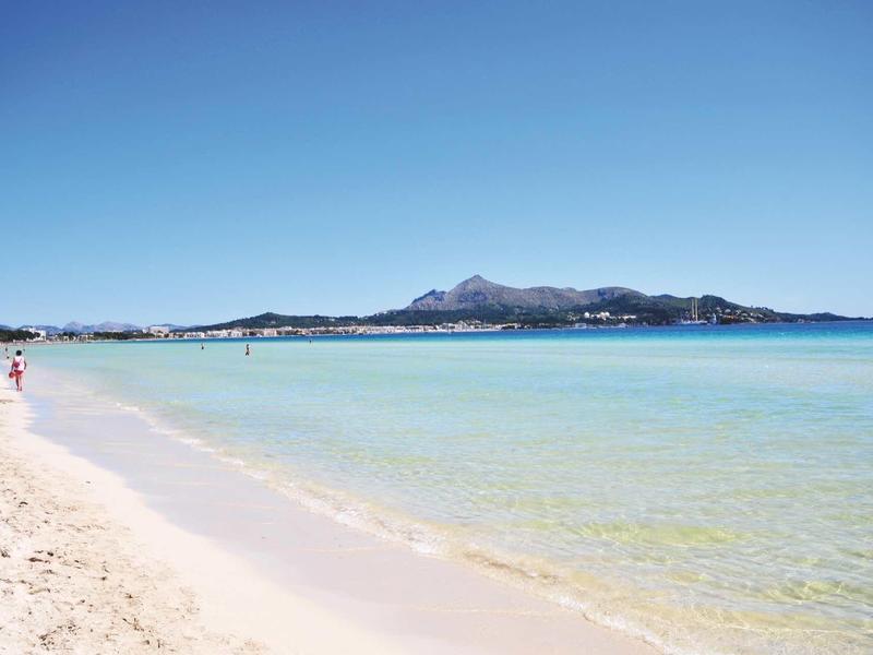 Jasná písčitá pláž s klidnou tyrkysovou vodou a horami v pozadí pod modrou oblohou.