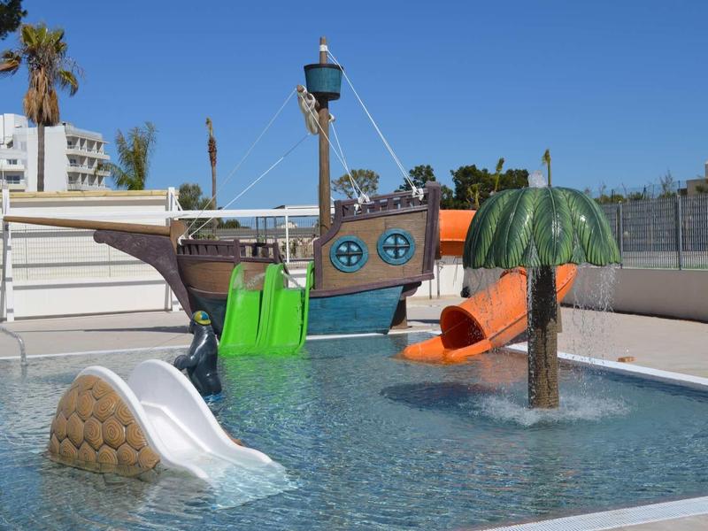 Malý venkovní bazén s barevným dětským hřištěm a skluzavkou pod modrou oblohou.