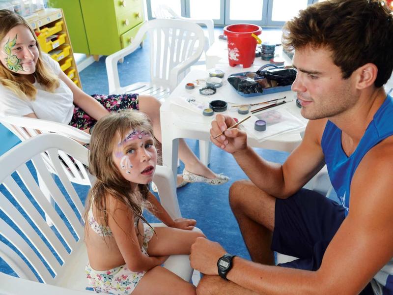 Muž maluje obličej malé dívce na dovolené, žena se dívá.