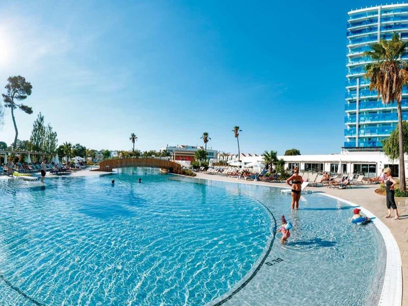 Velký venkovní bazén s čistou vodou, obklopený lehátky a palmami pod slunečnou oblohou.