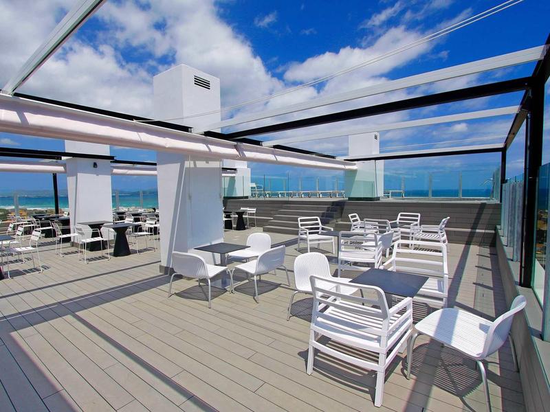 Moderní střešní terasa s bílými křesly, stoly a skleněným zábradlím pod modrou oblohou.