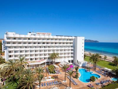Moderner Hotelkomplex mit Pool, Palmen und Meer im Hintergrund unter klarem Himmel.