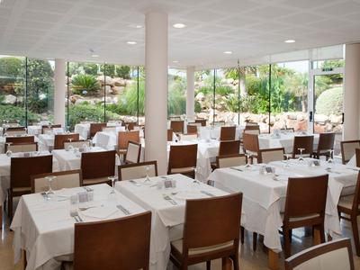 Helles Hotelrestaurant mit weißen Tischdecken und Holzmöbeln, umgeben von großen Fenstern mit Blick auf Garten.
