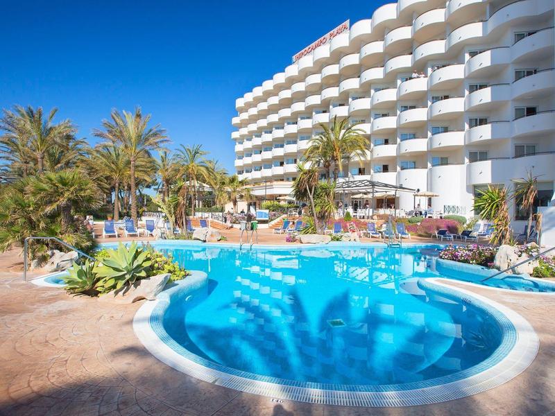 Hotel mit großem Pool, Palmen und blühenden Pflanzen unter klarem blauem Himmel.