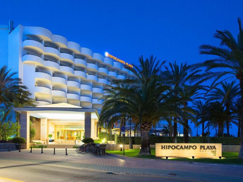 Modernes Hotelgebäude mit Palmen und beleuchtetem Eingang bei Dämmerung.