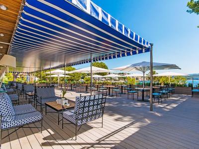 Terrasse avec tables et chaises, auvent bleu et vue sur la mer sous un soleil éclatant.