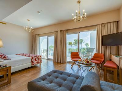 Chambre d'hôtel lumineuse avec deux lits, grandes fenêtres et coin salon avec chaises et téléviseur.