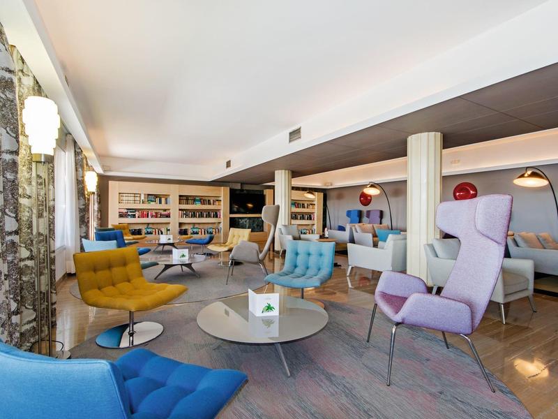 Salon d'hôtel moderne avec fauteuils colorés, tables basses et étagères dans une pièce lumineuse.